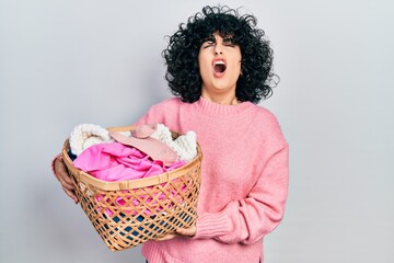 Young middle east woman holding laundry basket angry and mad screaming frustrated and furious,...