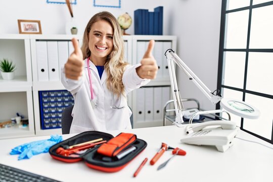 Young beautiful doctor woman with reflex hammer and medical instruments approving doing positive gesture with hand, thumbs up smiling and happy for success. winner gesture.