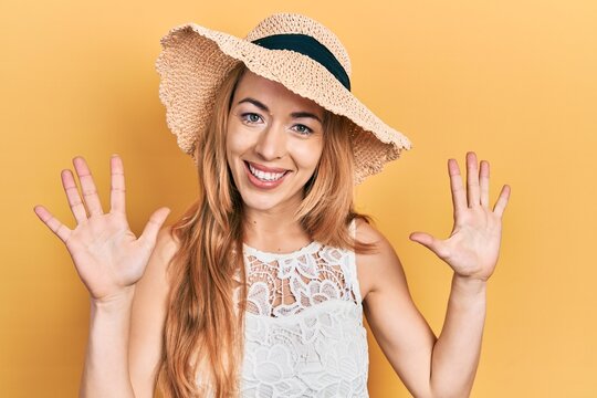 Young caucasian woman wearing summer hat showing and pointing up with fingers number ten while smiling confident and happy.