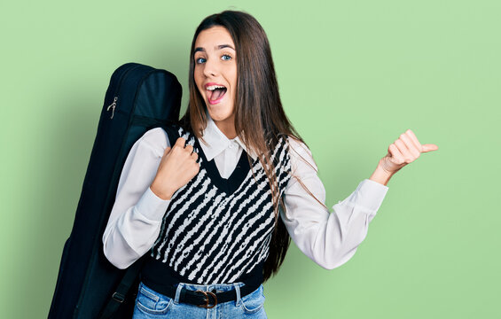 Young brunette teenager wearing guitar case pointing thumb up to the side smiling happy with open mouth