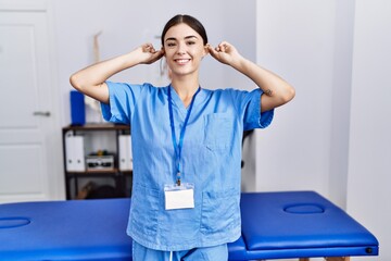 Young hispanic woman wearing physiotherapist uniform standing at clinic smiling pulling ears with fingers, funny gesture. audition problem