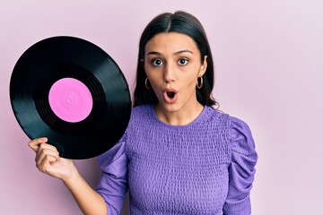 Young hispanic woman holding vinyl disc scared and amazed with open mouth for surprise, disbelief face