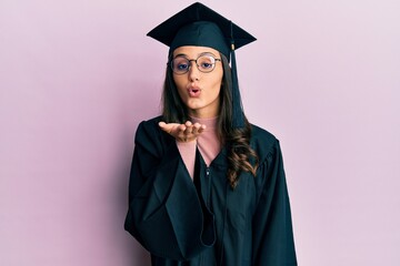 Young hispanic woman wearing graduation cap and ceremony robe looking at the camera blowing a kiss...