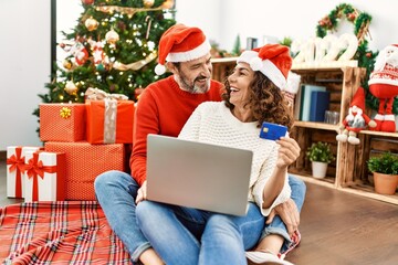 Middle age hispanic couple wearing christmas hat. Sitting on the floor using laptop and credit card at home.