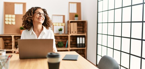 Middle age hispanic woman working at the office wearing glasses looking away to side with smile on face, natural expression. laughing confident.