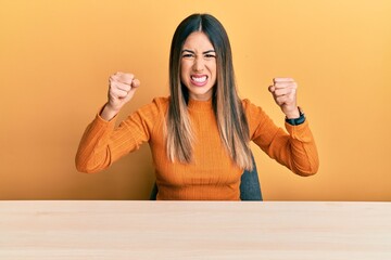 Young hispanic woman wearing casual clothes sitting on the table angry and mad raising fists...