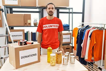 Young hispanic man wearing volunteer t shirt at donations stand winking looking at the camera with...
