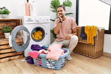 Young hispanic man putting dirty laundry into washing machine looking stressed and nervous with hands on mouth biting nails. anxiety problem.