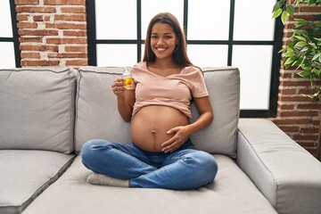 Young pregnant woman holding moisturizer oil to hydrate belly looking positive and happy standing and smiling with a confident smile showing teeth