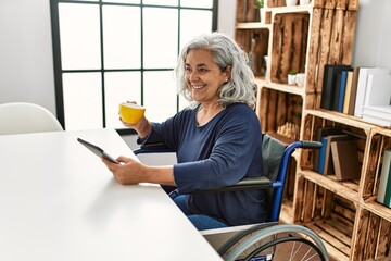 Middle age grey-haired disabled woman using touchpad and drinking cofffee sitting on wheelchair at home.