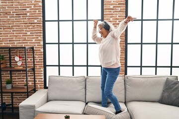 Middle age woman listening to music and dancing standing on sofa at home