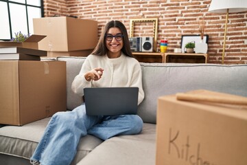 Young hispanic woman sitting on the sofa at new home using laptop with hand on head, headache...