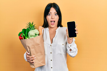 Young hispanic woman holding bag of groceries using smartphone afraid and shocked with surprise and...