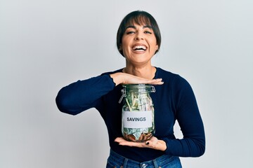 Young hispanic woman holding savings jar smiling and laughing hard out loud because funny crazy joke.