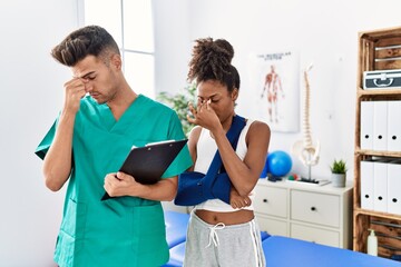 Physiotherapist working with patient wearing arm on sling at rehabilitation clinic tired rubbing...