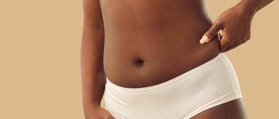 Fototapeta na wymiar Normalizing body flaws, accepting yourself and being confident in your imperfection: Plump young black woman in underwear, with a cute naked belly button, pinching some body fat on her side, close up