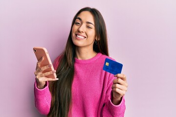 Young hispanic girl holding smartphone and credit card winking looking at the camera with sexy...