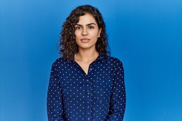 Young brunette woman with curly hair wearing casual clothes over blue background relaxed with...