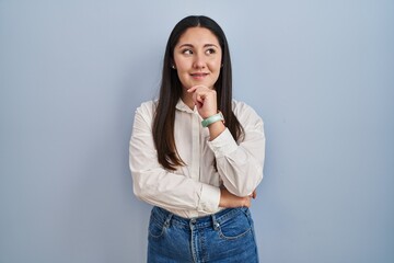 Young latin woman standing over blue background with hand on chin thinking about question, pensive expression. smiling and thoughtful face. doubt concept.