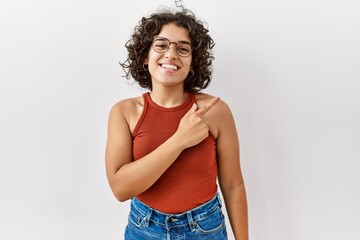 Young hispanic woman wearing glasses standing over isolated background cheerful with a smile on face pointing with hand and finger up to the side with happy and natural expression