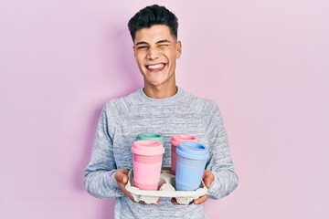Young hispanic man holding tray with take away coffee winking looking at the camera with sexy expression, cheerful and happy face.