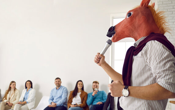 Bizarre speaker making funny absurd presentation in front of happy audience. Man wearing silly wacky horse mask holding microphone and giving lecture during business training, conference or seminar