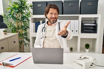 Handsome middle age doctor man working at the clinic smiling cheerful offering palm hand giving assistance and acceptance.