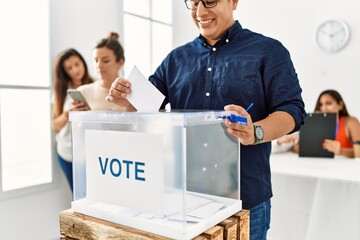 Young voter man smiling happy putting vote in voting box standing by ballot at electoral center.