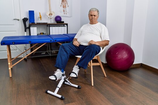 Senior Caucasian Man At Physiotherapy Clinic Using Pedal Exerciser Skeptic And Nervous, Frowning Upset Because Of Problem. Negative Person.