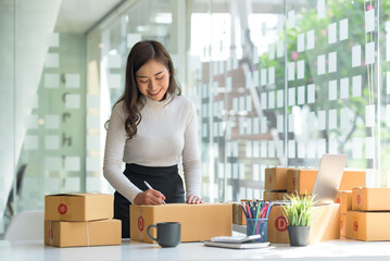 Smiling young Asian business owner woman prepare parcel box and standing check online orders of product for deliver to customer on laptop computer. Shopping Online concept.