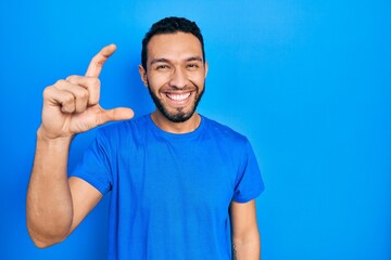 Fototapeta premium Hispanic man with beard wearing casual blue t shirt smiling and confident gesturing with hand doing small size sign with fingers looking and the camera. measure concept.