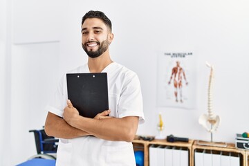 Young arab man wearing physiotherapist uniform holding clipboard at clinic - obrazy, fototapety, plakaty