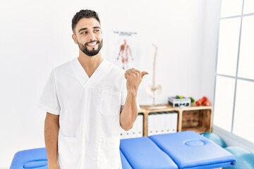 Young handsome man with beard working at pain recovery clinic smiling with happy face looking and pointing to the side with thumb up.