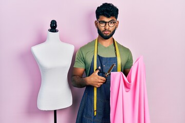 Arab man with beard dressmaker designer holding scissors and cloth skeptic and nervous, frowning...