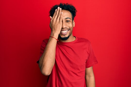 Young african american man with beard wearing casual red t shirt covering one eye with hand, confident smile on face and surprise emotion.