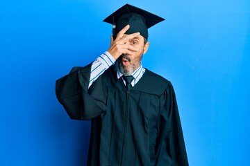 Middle age hispanic man wearing graduation cap and ceremony robe peeking in shock covering face and eyes with hand, looking through fingers with embarrassed expression.