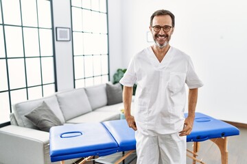 Middle age physiotherapy man working at home looking positive and happy standing and smiling with a confident smile showing teeth