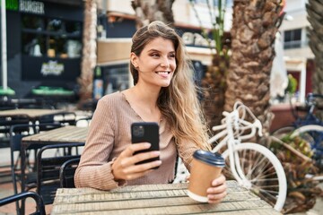 Young blonde woman using smartphone drinking coffee at coffee shop terrace