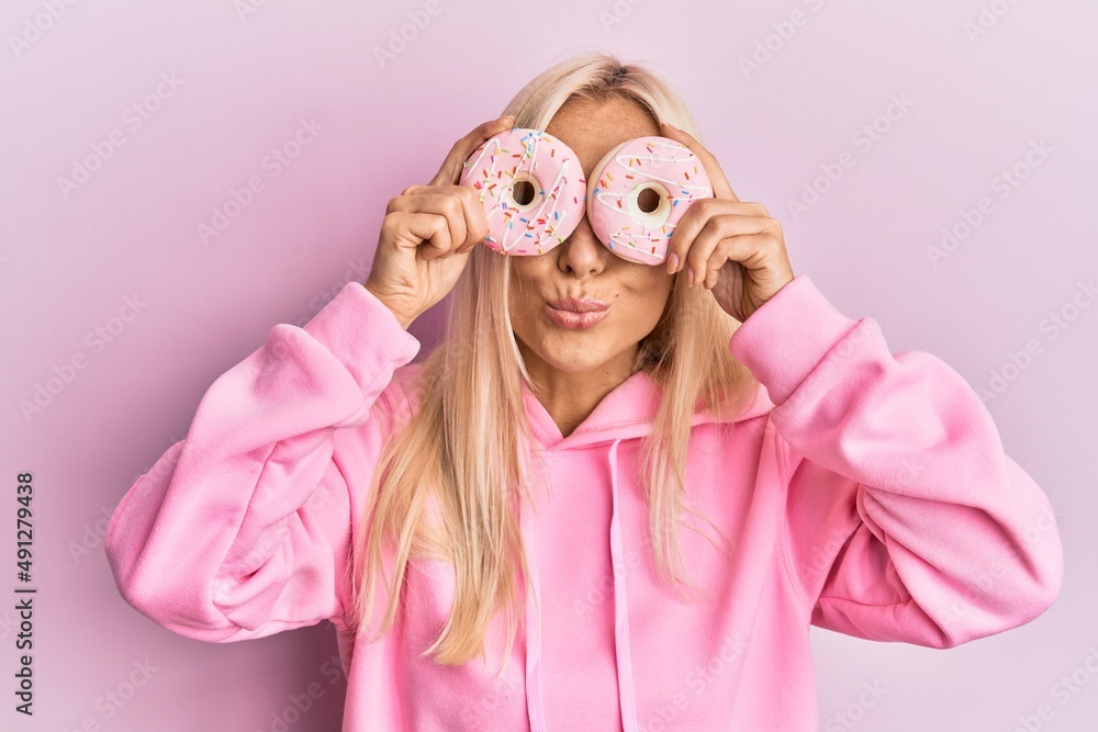 Canvas Prints young blonde woman holding tasty pink doughnuts over eyes looking at the camera blowing a kiss being