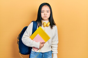 Young chinese girl holding student backpack and books puffing cheeks with funny face. mouth inflated with air, crazy expression.