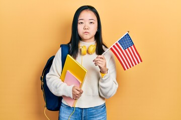 Young chinese girl exchange student holding america flag puffing cheeks with funny face. mouth inflated with air, catching air.