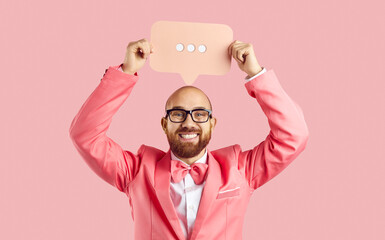 Positive funny man with social communication symbol in hands isolated on pastel pink background. Young man in pink suit and bow tie holds paper bubble over his head with three dots on it. Banner.