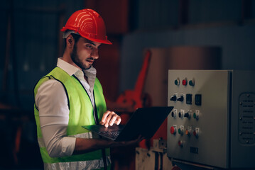 Factory worker checking electrical system and machine process via laptop on overtime job at...