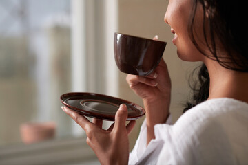 Cropped image of happy woman drinking cup of morning coffee and looking through apartment window in the morning