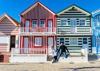 Fototapeta na wymiar Woman and man jumping for a photo in front of the typical colorful striped houses in Costa Nova, Portugal.