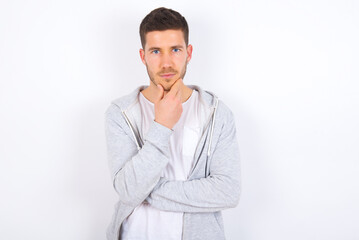 Thoughtful smiling young caucasian man wearing casual clothes over white background keeps hand under chin, looks directly at camera, listens something with interest. Youth concept.