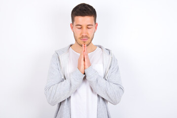 Indoor closeup of young caucasian man wearing casual clothes over white background practicing yoga and meditation, holding palms together in namaste, looking calm, relaxed and peaceful.