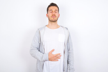 Satisfied smiling young caucasian man wearing casual clothes over white, keeps hands on belly, being in good mood after eating delicious supper, demonstrates she is full. Pleasant feeling in stomach.