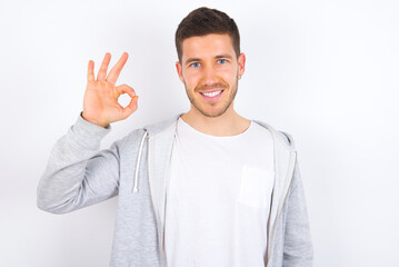 Glad attractive young caucasian man wearing casual clothes over white background shows ok sign with hand as expresses approval, has cheerful expression, being optimistic.