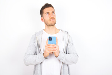 Portrait of young caucasian man wearing casual clothes over white background  with dreamy look, thinking while holding smartphone. Tries to write up a message.